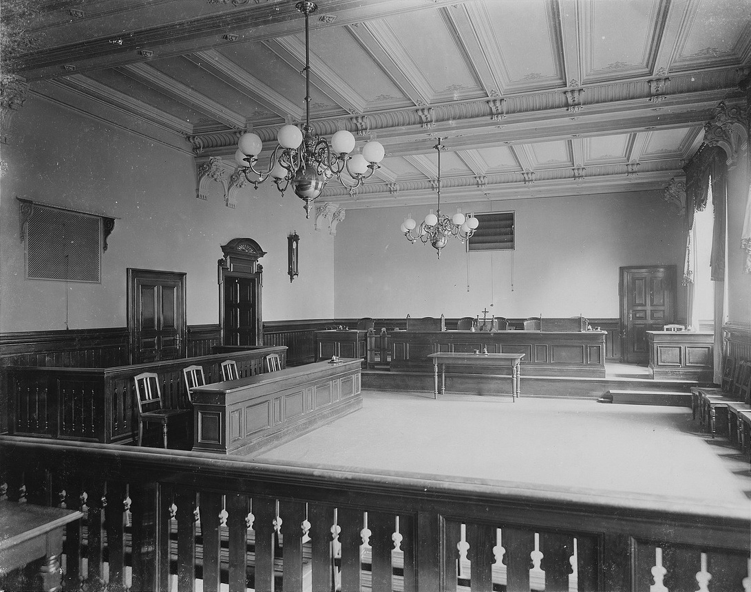 Ein Gerichtssaal mit zwei Kronleuchtern, Tischbänken und Sesseln aus Holz.  © Wikimedia