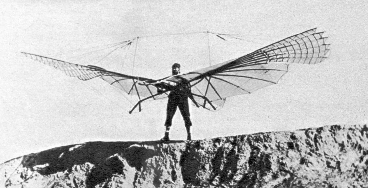 Ein Mann steht auf einer Felsklippe. Er trägt einen Flugapparat, der aussieht wie zwei Flügel, die mit Seilen und Drähten miteinander verbunden sind. © Wikimedia