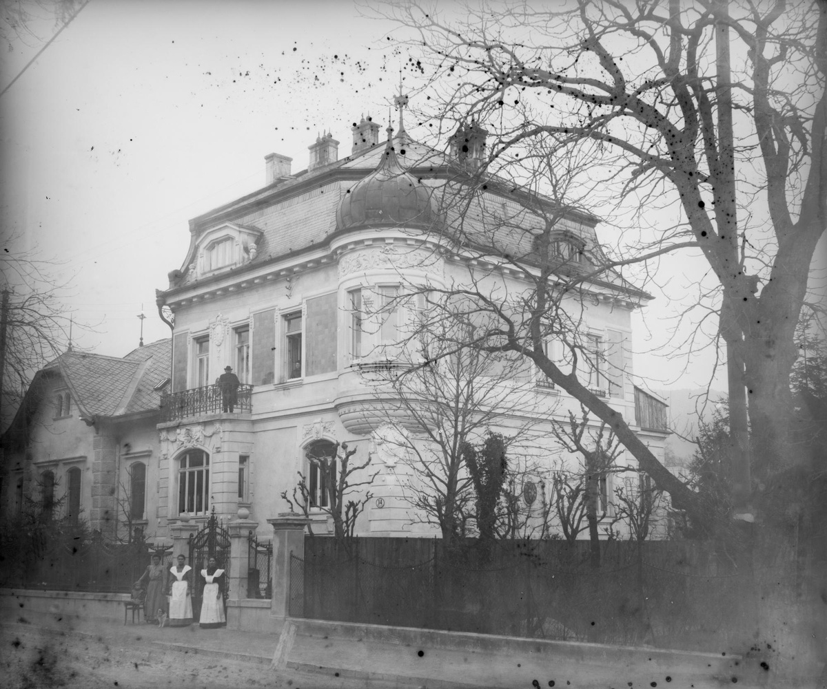 Zwei Dienstmädchen und eine Hausherrin vor dem Gartentor einer herrschaftlichen Villa mit Erker und Balkon, rechts ein großer Baum, links hinten ein weiteres Haus. © ÖNB Bildarchiv