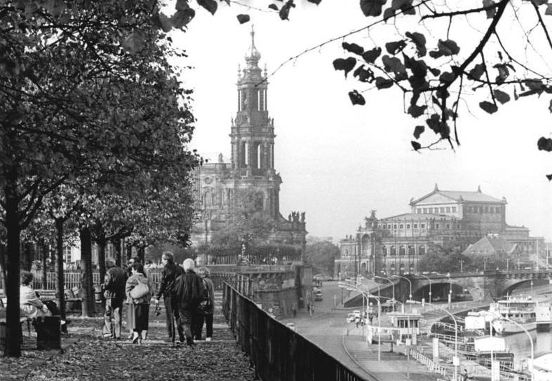 Schwarzweißfotografie einer Straßenansicht in Dresden mit Semperoper und Hofkirche. Links stehen Bäume. Einige Passanten gehen einen Wenig entlang. Rechts ist eine Straße.  © Klaus Oberst, Wikimedia, CC-BY-SA-3.0-DE. 