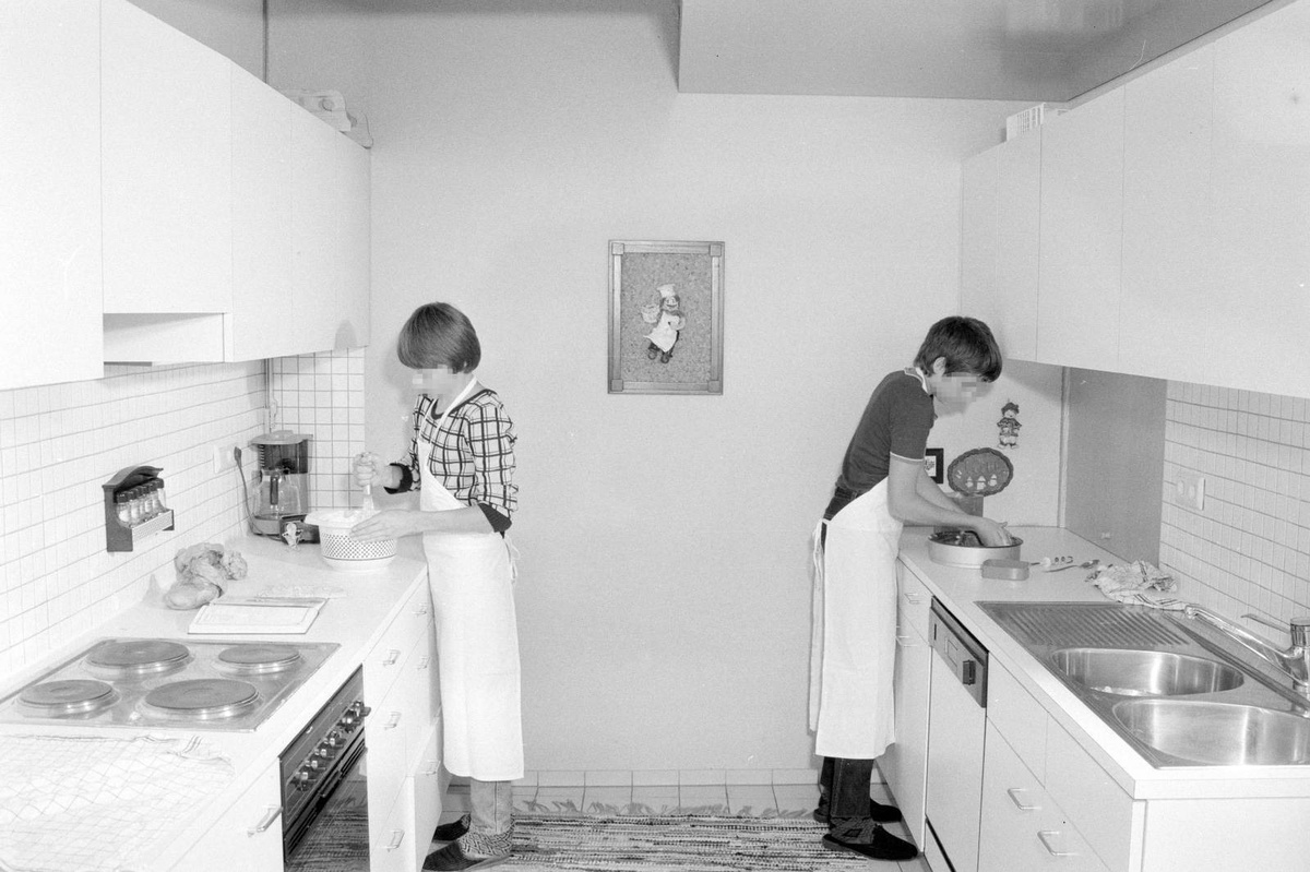 Ein Foto aus den Innenräumen des Heims. Zwei Jugendliche stehen in einer kargen, vollkommen weißen Küche. Die Jungen tragen weiße Schürzen. Der linke rührt mit einem Schneebesen einen Teig an. Der rechte fettet eine Teigform ein. Zwischen beiden hängt das einfach gezeichnete Bild eines Kochs an der Wand. © Vorarlberger Landesbibliothek, Helmut Klapper, Jugendheim Jagdberg, 1985.