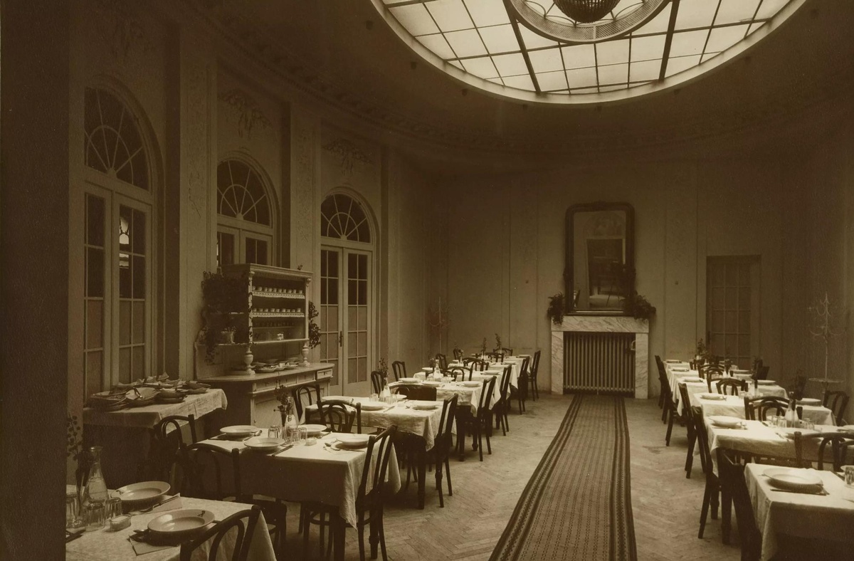 Speisesaal mit zwei Reihen mit gedeckten Tischen und Stühlen, am Ende des Zimmers ein Kamin, links ein Kasten und große Glastüren. © Wien Museum