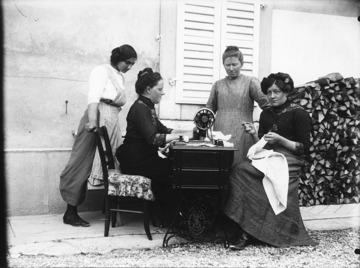 Vier Frauen sitzen und stehen um eine Nähmaschine. Eine Frau näht an der Maschine, eine andere von Hand.  © Wikimedia