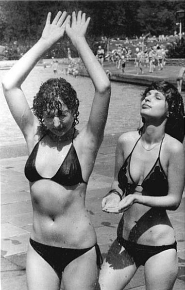 Zwei Frauen im Bikini duschen in einem Freibad. Eine hält die Arme nach oben, wodurch ihre rasierten Achseln freiliegen. Im Hintergrund befinden sich ein Pool und Freibadbesucher:innen.  © Wikimedia, Bundesarchiv, Bild 183-1982-0515-015, Klaus Franke, Frauen in einem Berliner Freibad, 1982, CC-BY-SA 3.0