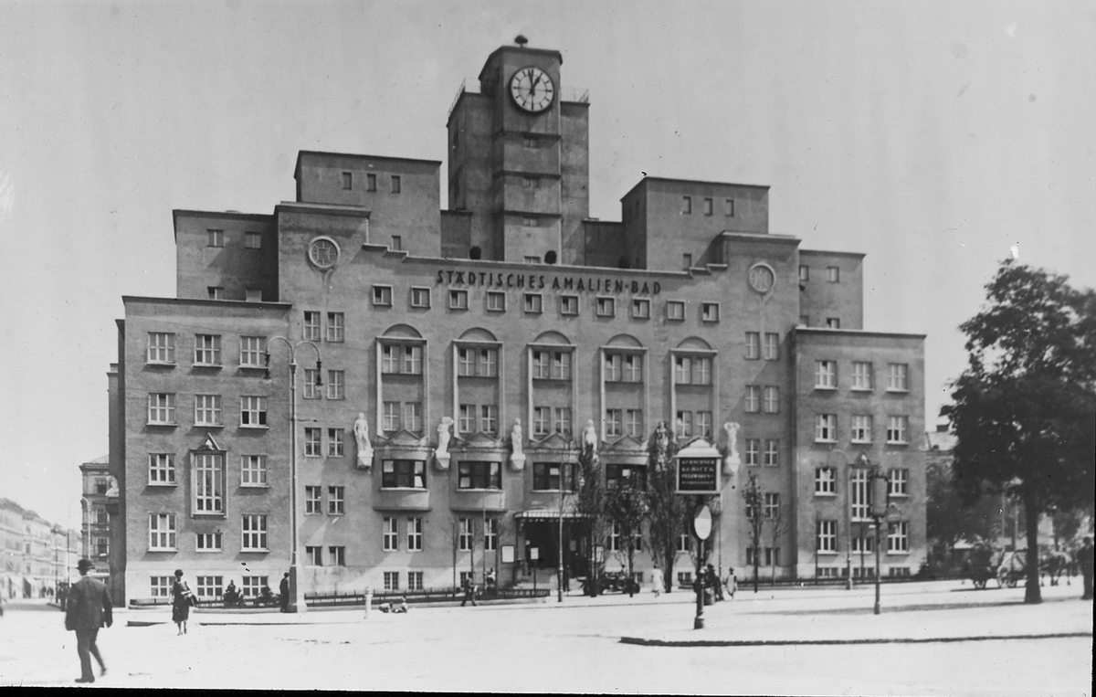 Das Foto zeigt das Amalienbad. Die Fassade erscheint auf dem Bild hellgrau. Die verschiedenen Gebäudeteile sind unterschiedlich hoch und symmetrisch angeordnet. In der Mitte ragt ein breiter Turm auf, an dessen Vorderseite sich eine Uhr mit weißem Zifferblatt befindet. Das große Gebäude zieren an der Fassade sechs weiße Statuen. Weiter oben befindet sich der Schriftzug „Städtisches Amalien-Bad“. Auf dem Platz vor dem Gebäude sind einige Fußgänger:innen unterwegs. Rechts vom Bad steht ein Baum.  © Wikimedia, ETH-BIBLIOTHEK, Amalienbad, um 1925 (CC-BY-SA-4.0). 