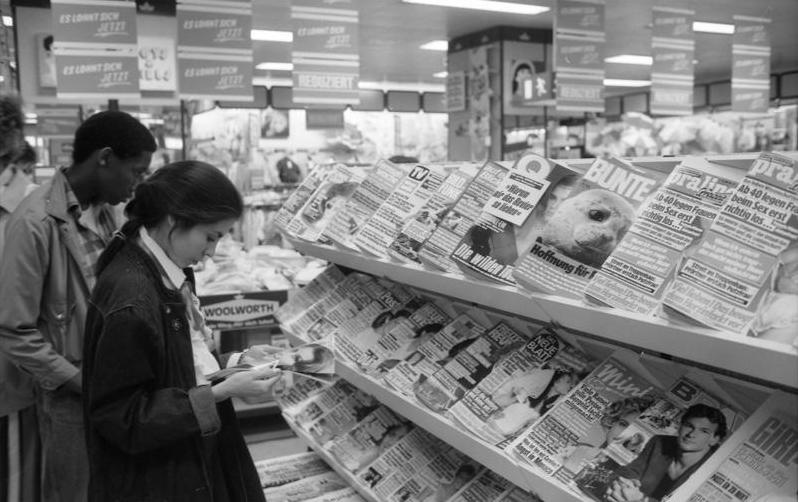 Ein Junge und ein Mädchen stehen in einem Geschäft vor Regalen mit Zeitschriften. Das Mädchen blättert in einer Zeitschrift. Rechts unten ist die BRAVO im Regal zu sehen.  © Wikimedia, Bundesarchiv, B 145 Bild-F079046-0031, Geschäft in Bad Godesberg mit Zeitschriften, 1988 (CC-BY-SA 3.0). 