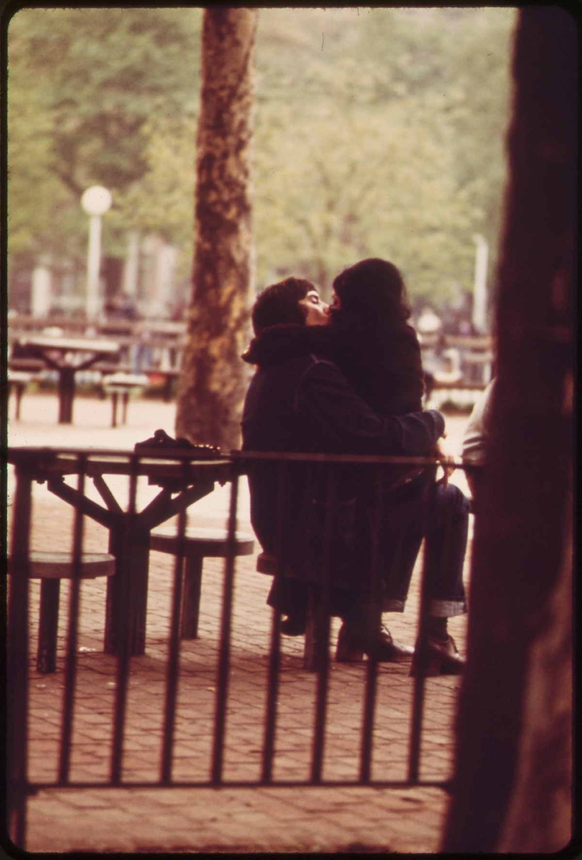 Ein Mann und eine Frau sitzen umschlungen auf einer Bank und küssen sich. Im Hintergrund befinden sich Bänke und ein Park.  © Wikimedia, küssendes Paar, New York, 1973. 
