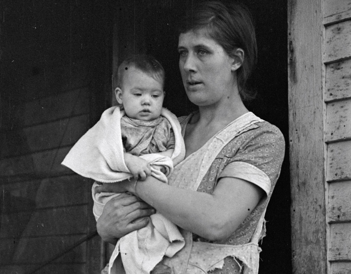 Eine Mutter mit dunklen Haaren hält ein Baby in den Armen. Sie trägt ein Kleid und darüber eine zerrissene Schürze. Das Baby ist in ein Tuch gewickelt.  © Wikimedia, Frau Atkins mit Baby, USA, 1936. 