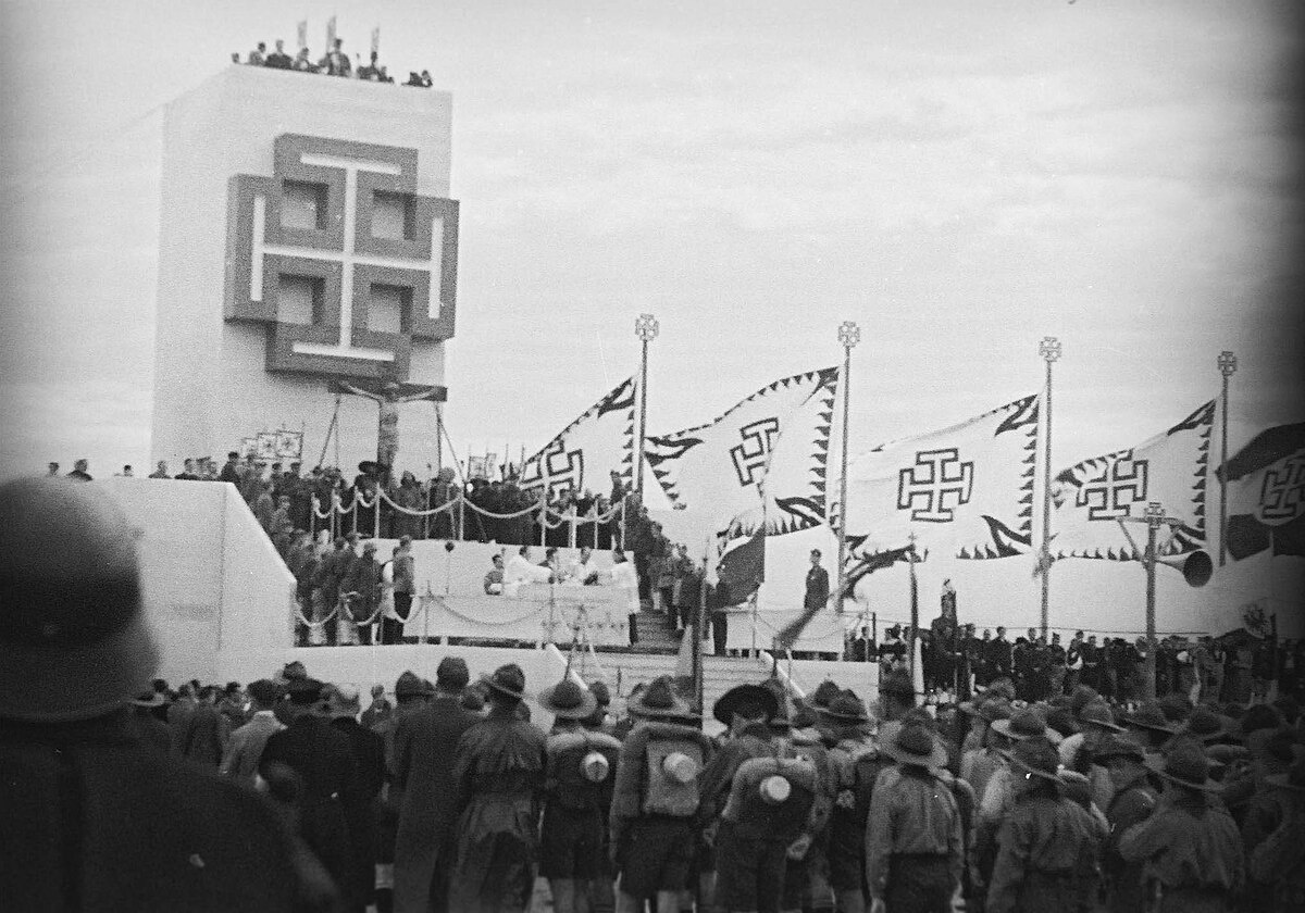 Das Foto zeigt eine Veranstaltung der Austrofaschisten. Auf einem Turm ist ein riesiges Kruckenkreuz zu sehen, davor befindet sich ein Kreuz mit Jesusfigur auf einer Tribüne. Rechts neben dem Turm sind mehrere große Fahnen mit Kruckenkreuzen. Auf und vor der Tribüne stehen viele Menschen.  © Wikimedia, Propagandaveranstaltung im Austrofaschismus, 1936. 
