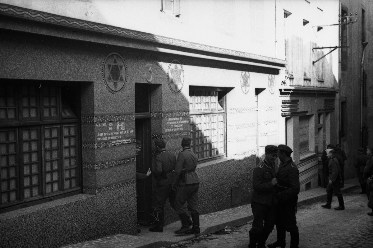 Zwei Wehrmachtssoldaten betreten ein Bordell. Durch Davidsterne an der Fassade ist das Gebäude als ehemalige Synagoge erkennbar. Auf der Straße vor dem Gebäude halten sich einige weitere Soldaten auf.  © Wikimedia, Bundesarchiv, Bild 101II-MW-1019-07/Dietrich, Wehrmachtsbordell, Frankreich, 1940 (CC-BY-SA 3.0). 