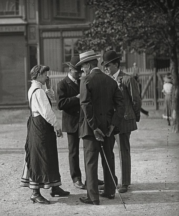 Drei Männer mit Anzug und Hut, einer mit Stock, stehen auf einer ungepflasterten Straße, neben ihnen eine Frau mit Flechtfrisur, langem Kleid und Schürze. Im Hintergrund ein Gartenzaun, ein Baum und ein Haus. © Wikimedia