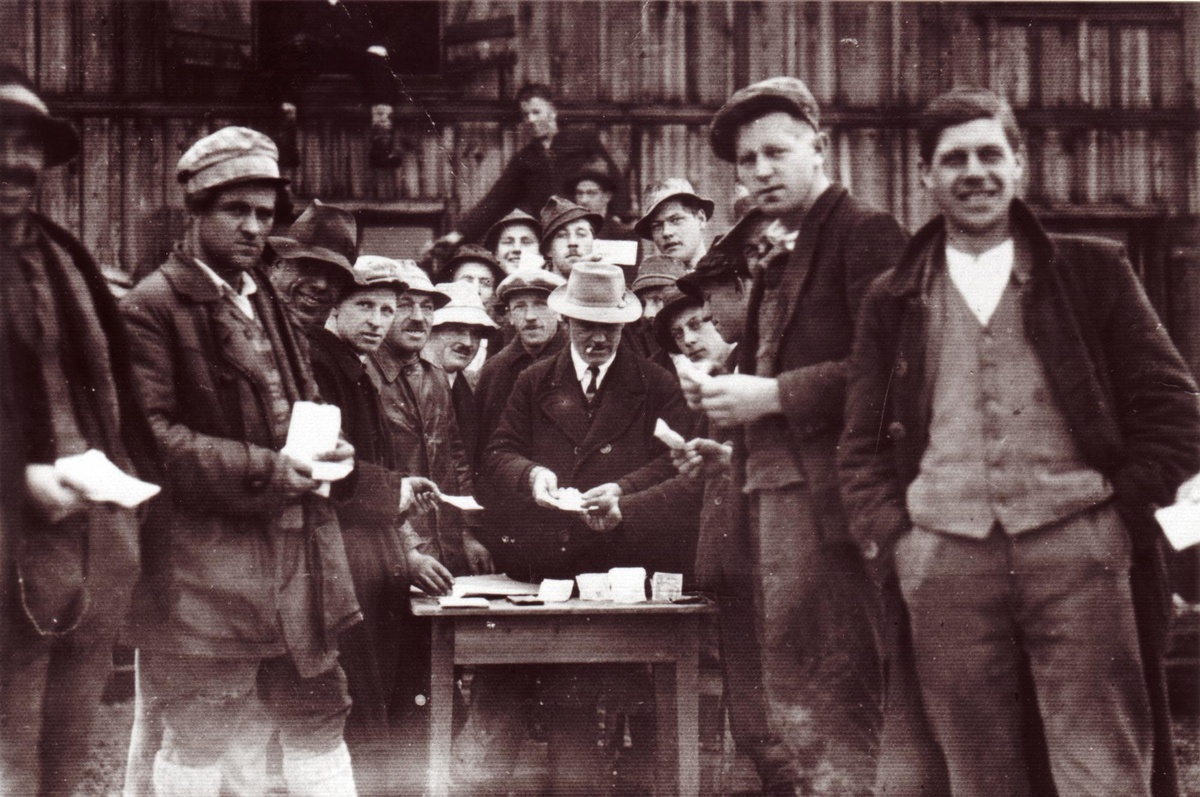 Eine Gruppe von Arbeitern, teilweise mit Hüten und Kappen, steht um einen Tisch, wo der Arbeitgeber mit Hut und Zigarette im Mund Scheine zählt und ausgibt. © Unterguggenberger Institut 