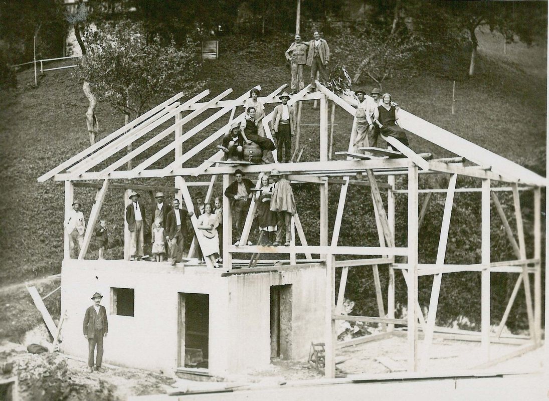 Männer und Frauen in festlicher Kleidung sitzen und stehen auf dem Holzgerüst eines Hauses, das noch nicht fertig gebaut wurde. © Unterguggenberger Institut 