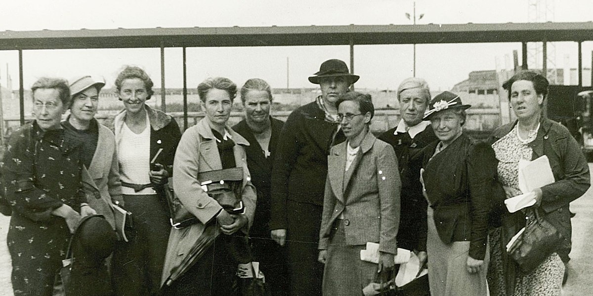 Eine Gruppe Frauen in festlicher Kleidung, einige mit Hut und Blazer, stehen nebeneinander und blicken in die Kamera. Dahinter ein Bahnsteig und eine Wiese.  © Wikimedia