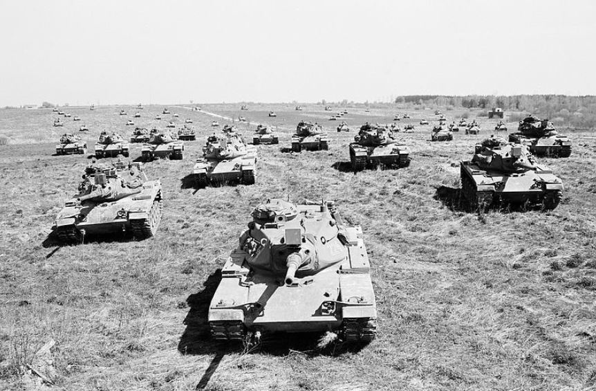 Viele Panzer stehen auf einer großen Wiese. Auf der rechten Seite ist im Hintergrund Wald zu sehen.  © Wikimedia, Bundesheerfotos, Panzer bei einer Übung am Truppenübungsplatz Döllersheim-Allensteig, 1987 (CC-BY-SA-2.0). 