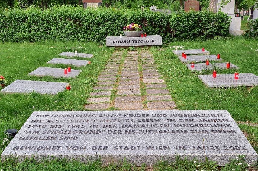 Auf einer Wiese mit einem Gehweg aus Platten befinden sich mehrere Grabsteine mit roten Kerzen. Im Hintergrund vor einer Hecke steht auf einem Stein der Schriftzug „Niemals vergessen“. Im Vordergrund steht auf einer Steinplatte: „Zur Erinnerung an die Kinder und Jugendlichen, die als ‚lebensunwertes Leben‘ in den Jahren 1940 bis 1945 in der damaligen Kinderklinik ‚am Spiegelgrund‘ der NS-Euthanasie zum Opfer gefallen sind. Gewidmet von der Stadt Wien im Jahre 2002.“ © Wikimedia, Haeferl, Gedenktafel für die am Spiegelgrund ermordeten Kinder am Zentralfriedhof, 2015 (CC-BY-SA-3.0). 