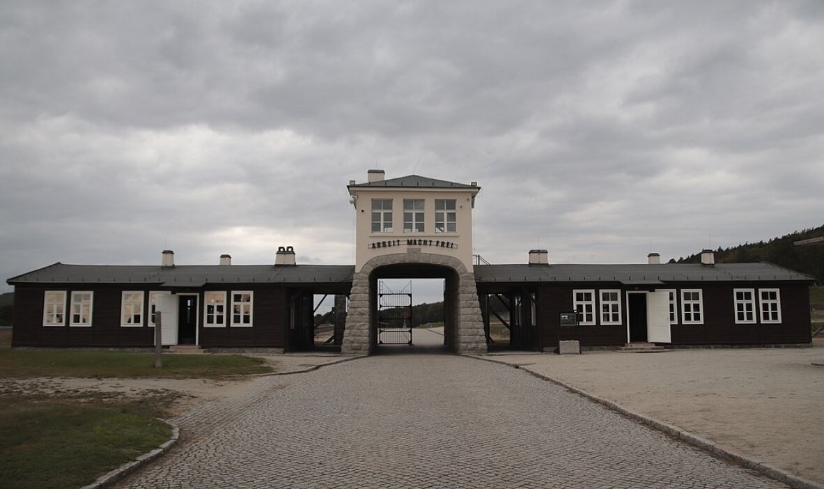 Ein Tor mit der Aufschrift „Arbeit macht frei“ wird auf beiden Seiten von niedrigeren Bauten flankiert. Eine gepflasterte Straße führt auf das Tor zu. Der Himmel ist bewölkt.  © Wikimedia, Gordon Roemhild, Eingangstor des KZ Groß-Rosen, CC-BY-SA-4.0. 