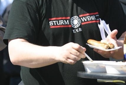 Das Bild zeigt den Oberkörper eines Mannes, der Würste von einem Pappteller isst. Er trägt ein schwarzes T-Shirt mit der Aufschrift „STURMWEHR T.Y.R“. In der Mitte des Shirts befindet sich das Symbol „Schwarze Sonne“, das in der rechtsextremen Szene beliebt ist.  © Wikimedia, T-Shirt mit „Schwarzer Sonne“ sowie der Abkürzung T.Y.R. (Tyr), vor 2009, (GFDL 1.2). 