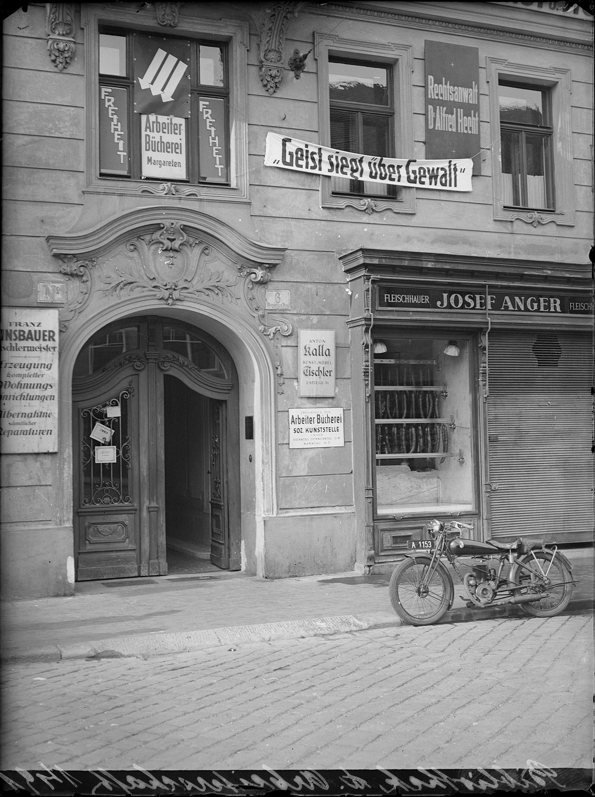 Das Bild zeigt die Arbeiterbücherei Margareten. An der Fassade hängen mehrere Schilder und Transparente, eines davon mit der Aufschrift “Geist siegt über Gewalt”. Vor der Bücherei steht am Straßenrand ein Motorrad.  © ÖNB Bildarchiv, Arbeiterbücherei Margareten, 1933. 