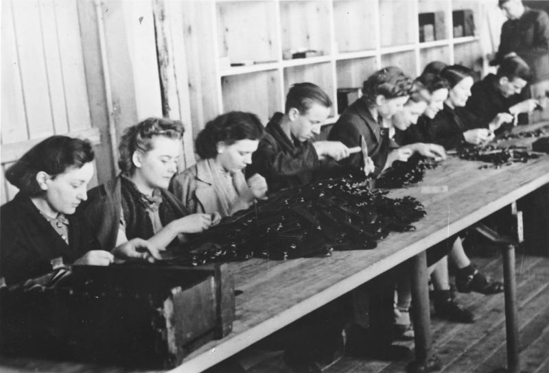 Arbeiter:innen sitzen an einem langen Tisch in einer Reihe und verrichten mit den Händen verschiedene Arbeiten. Sie haben den Kopf nach vorne geneigt.  © Wikimedia, Bundesarchiv , Bild 183-L12005, Zwangsarbeiter:innen in einer Gewehrfabrik in Polen (CC-BY-SA 3.0). 