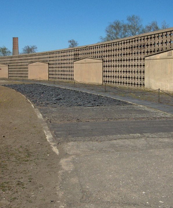 Hinter einer Laufstrecke aus unterschiedlichen Materialien wie Beton, Steinen und Sand steht eine hohe Mauer. Im Hintergrund sieht man einen Schornstein. © Wikimedia, Ukas, Teststrecke Schuhläuferkommando, 2005.