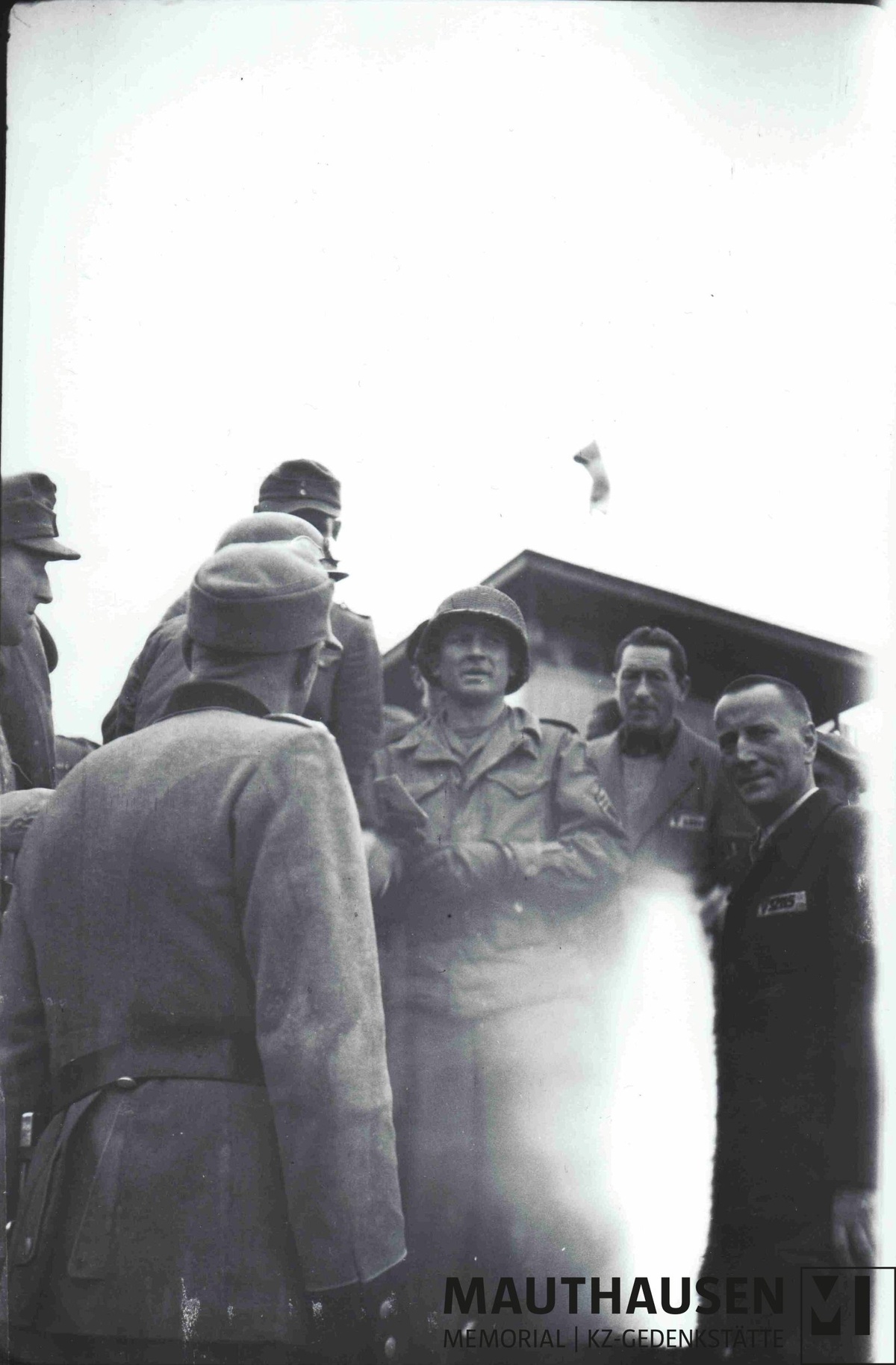 In der Mitte des Schwarz-Wei&szlig;-Fotos befindet sich Albert Kosiek. Er tr&auml;gt Helm und Milit&auml;runiform. Rechts im Bild befinden sich &uuml;berlebende H&auml;ftlinge mit H&auml;ftlingsnummern auf der Brust. Links im Bild stehen Feuerschutzpolizisten in Uniform.  Unten rechts befindet sich ein wei&szlig;er Fleck durch eine Besch&auml;digung der Kamera. &copy; Mauthausen Memorial, Sammlung Antonio Garcia, Fotograf: Francisco Boix, S 4665, Kosiek mit Wachen der Feuerschutzpolizei, 5. Mai 1945, Signatur: 4.2.1.006.B3.