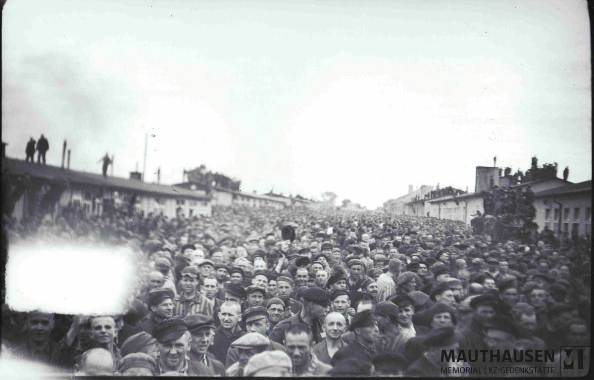 Das Schwarz-Wei&szlig;-Foto zeigt eine gro&szlig;e Menge von KZ-H&auml;ftlingen auf dem Appellplatz. Die meisten schauen in Richtung der Kamera. Einige tragen noch die gestreifte H&auml;ftlingskleidung. Rechts und links des gro&szlig;en Platzes sind Baracken zu sehen. Manche H&auml;ftlinge stehen auf den D&auml;chern der Baracken. Links im Bild befindet sich ein wei&szlig;er Fleck durch eine Besch&auml;digung der Kamera. &copy; Mauthausen Memorial, Sammlung Antonio Garcia, S 4665, Fotograf: Francisco Boix, H&auml;ftlinge auf dem Appellplatz warten auf die US-Soldaten, 5. Mai 1945, Signatur: 4.2.2.002.B2.