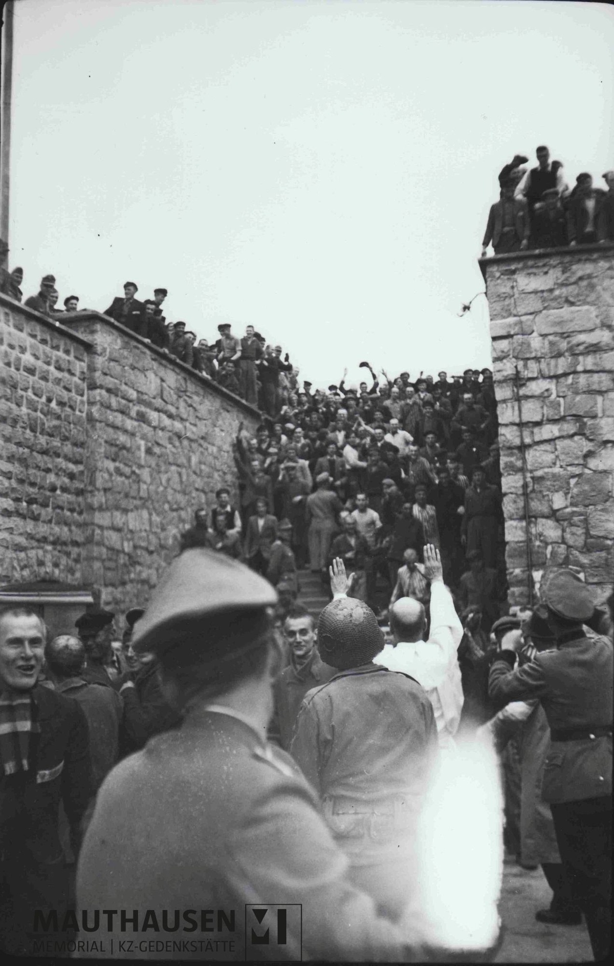Im Vordergrund des Schwarz-Wei&szlig;-Fotos befindet sich ein Feuerschutzpolizist in Uniform und mit M&uuml;tze, der mit dem R&uuml;cken zur Kamera steht. In der Bildmitte steht Albert Kosiek mit Helm, ebenfalls mit dem R&uuml;cken zur Kamera. Vor ihm hebt ein Mann in wei&szlig;em Kittel, vermutlich ein H&auml;ftlingsarzt, beide Arme nach oben. Im Hintergrund befindet sich eine Treppe zwischen zwei Mauern. Auf der Treppe und den Mauern stehen zahlreiche H&auml;ftlinge, einige davon mit jubelnd erhobenen Armen. &copy; Mauthausen Memorial, Sammlung Antonio Garcia, Fotograf: Francisco Boix, S 4665, Albert Kosiek im Garagenhof mit &Uuml;berlebenden und Wachen der Feuerschutzpolizei, 5. Mai 1945, Signatur: 4.2.2.006.B1.