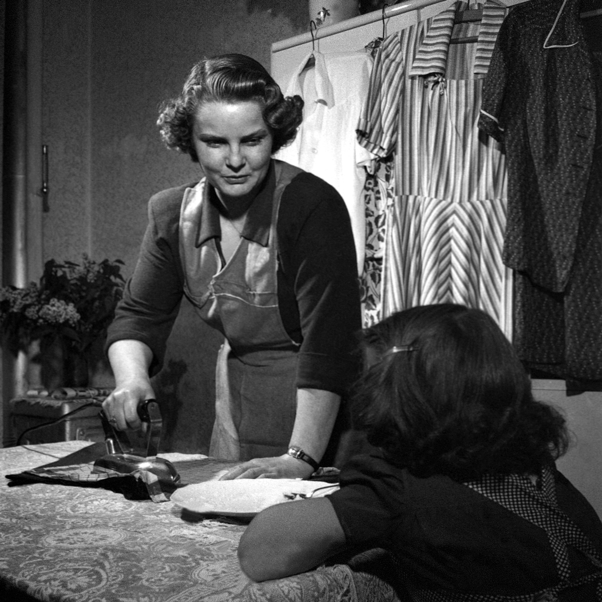 Eine Frau mit gewellten Haaren, Bluse und Schürze bügelt am Esstisch. Sie spricht zu einem Kind, das vor ihr sitzt. Im Hintergrund Kleidungsstücke auf Kleiderbügeln und eine Blumenvase. © ÖNB Bildarchiv, Hausfrau, 1950. 