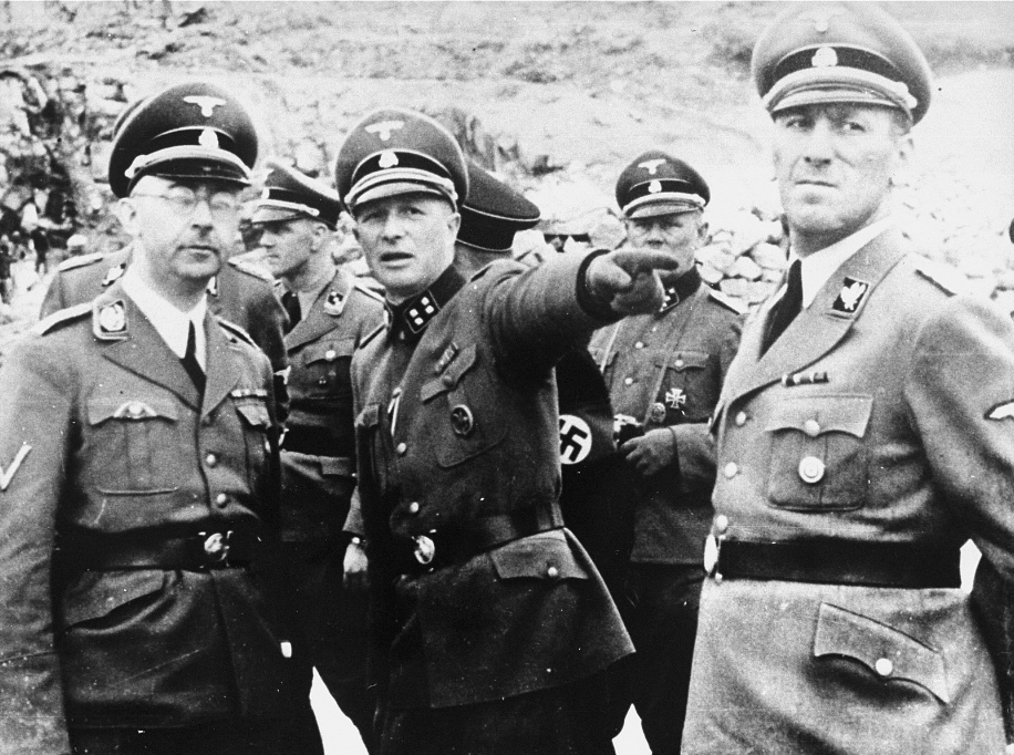 Das Schwarz-Wei&szlig;-Foto zeigt Heinrich Himmler, Lagerkommandant Franz Ziereis und Obergruppenf&uuml;hrer Ernst Kaltenbrunner im Steinbruch. Sie tragen SS-Uniformen. Franz Ziereis hat seinen Arm nach vorne ausgestreckt und zeigt auf etwas au&szlig;erhalb des Bildes. Im Hintergrund befinden sich weitere SS-Angeh&ouml;rige und H&auml;ftlinge im Steinbruch. &copy; United States Holocaust Memorial Museum, Foto des Erkennungsdiensts im KZ Mauthausen, Heinrich Himmler, Franz Ziereis und Ernst Kaltenbrunner beim Besuch im KZ, April 1941, gezeigt bei Boix&lsquo; Zeugenaussage in N&uuml;rnberg, Signatur: 12059. 