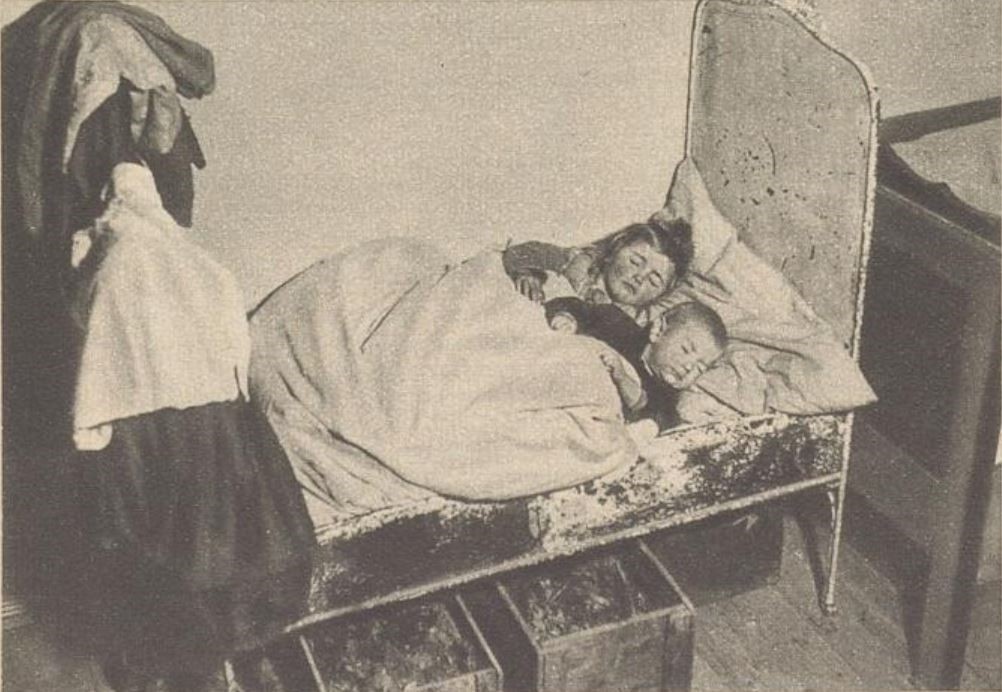 Fotos von Arbeiterfamilien in Grünbach: Zwei Kinder schlafen in einem kleinen Metallbett, eine Familie sitzt am Esstisch. © Anno