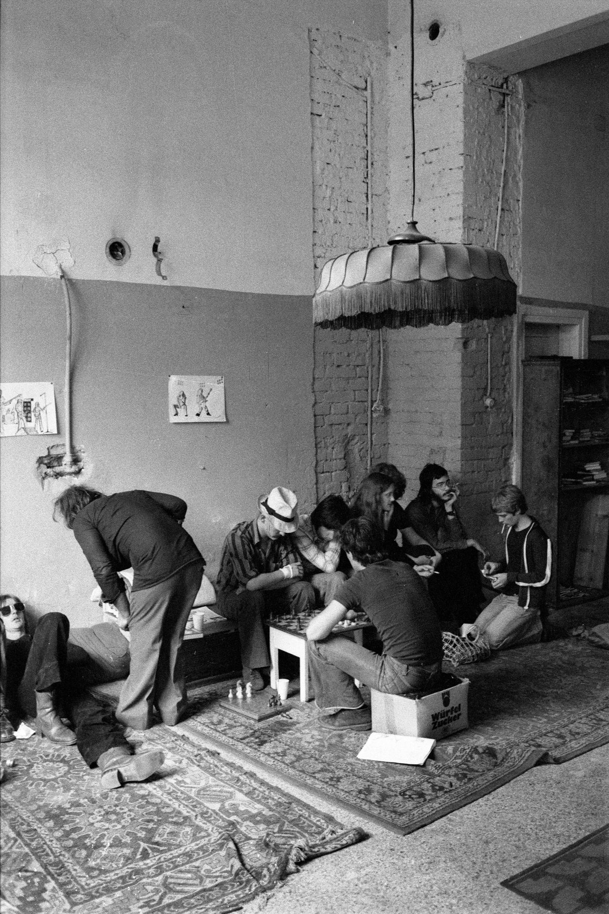 In einem Innenraum der Arena sitzen und liegen junge Menschen am Boden und auf Bierkisten. Der Boden ist mit Teppichen ausgelegt. Einige spielen Schach oder rauchen. Von der Decke hängt ein großer Lampenschirm.  © Heinz Riedler, Wien Museum. 