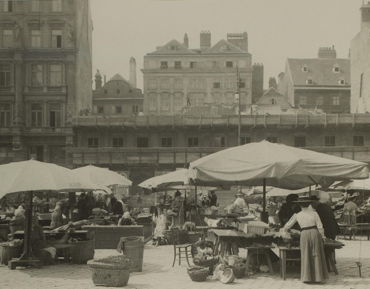 Marktplatz mit Ständen und Schirmen, rechts vorne steht eine Frau und greift zur Ware. Dahinter eine hohe Häuserfront. © Wien Museum
