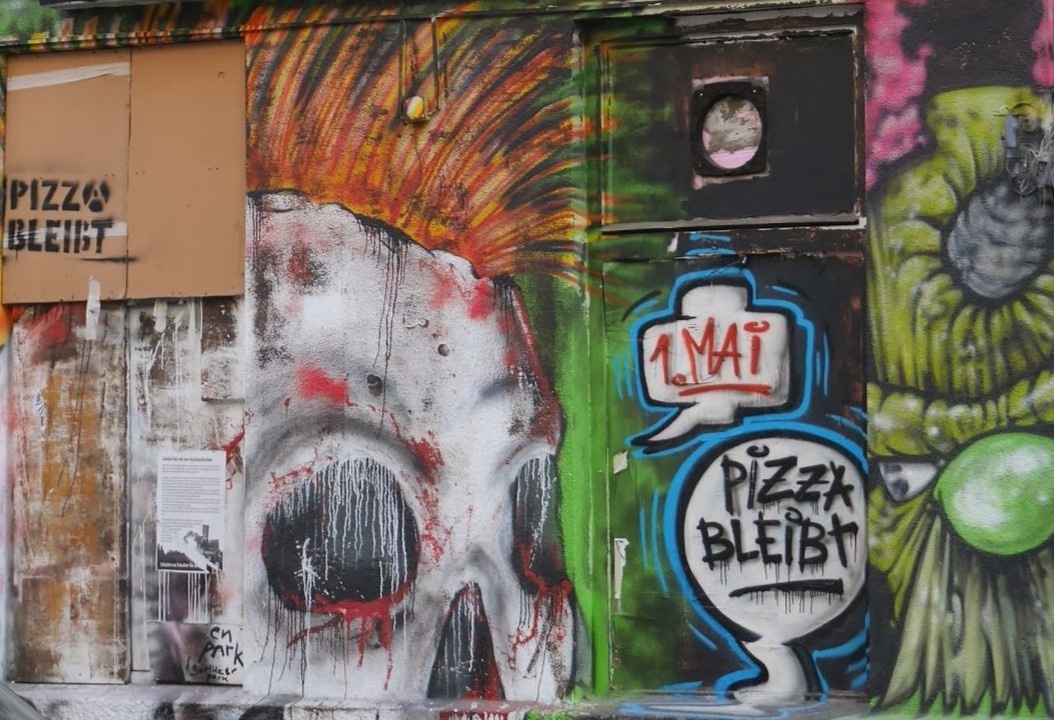 Das Bild zeigt Graffiti auf der Fassade des besetzten Hauses. Links befindet sich ein Totenkopf in weißer und roter Farbe, rechts befinden sich zwei Sprechblasen mit dem Text „1. Mai“ und „Pizza bleibt“. Auch auf einem Pappkarton, der oben links im Bild an der Fassade angebracht ist, ist der Slogan „Pizza bleibt“ zu lesen.  © Wikimedia, Harald Schilly, Fassade der Mühlfeldgasse 12 (CC-BY-SA-3.0). 