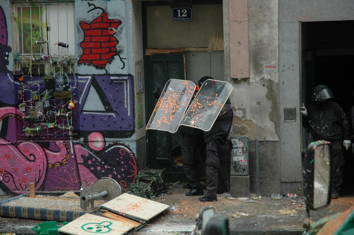 Vor der Fassade des besetzten Hauses stehen zwei Polizist:innen, die ihre Schilder über dem Kopf halten. Auf den Schildern befinden sich Farbspritzer. Das Haus ist mit Graffiti bemalt.  © Wikimedia, Otto Normalverbraucher, Räumung der Mühlfeldgasse 12, 2014 (CC-BY-SA-4.0). 