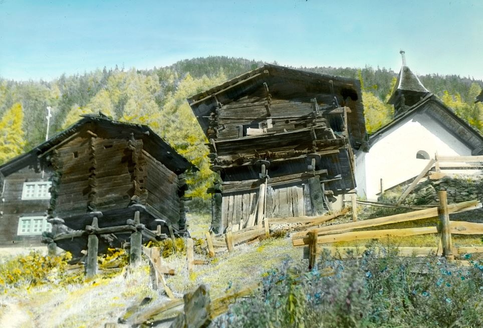 Kleine, windschiefe Häuser aus dunkelbraunem Holz und eine kleine Dorfkirche stehen vor einem waldigen Berg. © Wikimedia, Leo Wehrli, ETH Library.  
