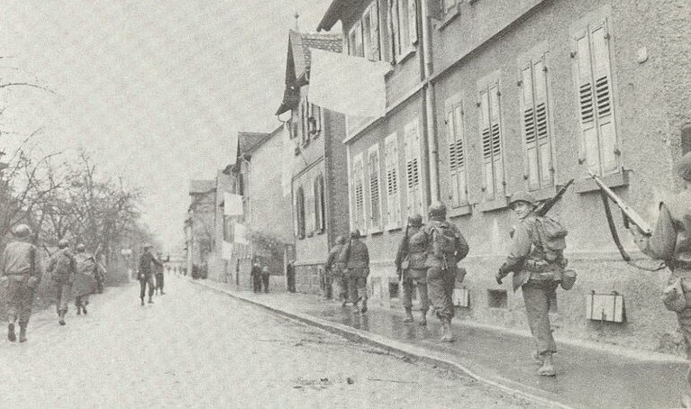 Soldaten gehen auf einer Straße an einer Häuserzeile entlang. An einem Fenster weht eine weiße Fahne.  © Wikimedia, weiße Fahnen beim Durchmarsch von US-Soldaten im Rheinland, 1945. 