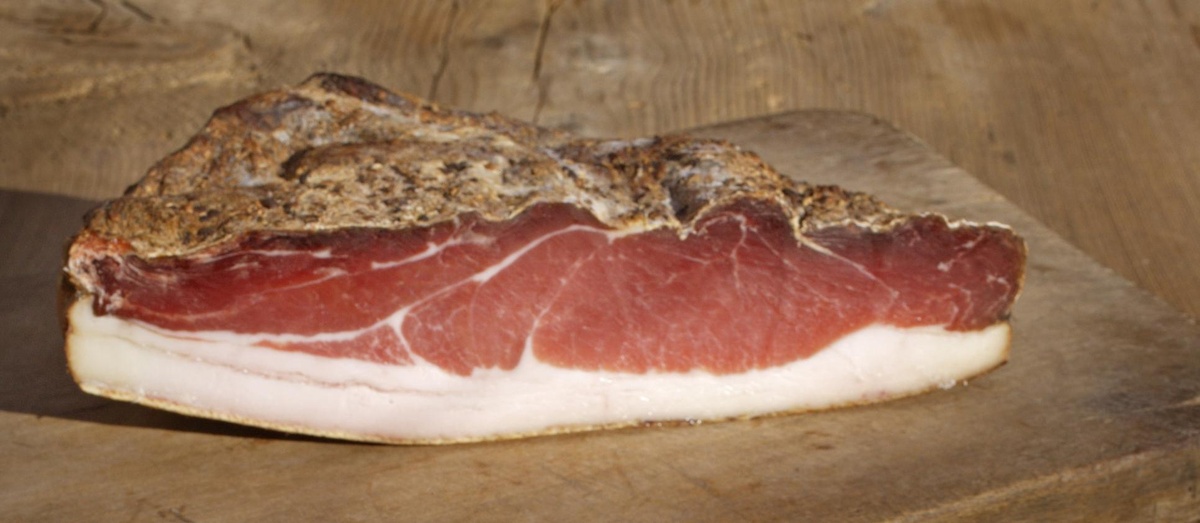 Auf einem Holzstück liegt ein Holzbrett mit einem großen Stück Speck darauf. Der Speck hat auf der Unterseite eine weiße Speckschwarte.  © Wikimedia, Fotograf Frieder Blickle, Südtiroler Speck g.g.A., 2010 (CC-BY-SA-4.0). 