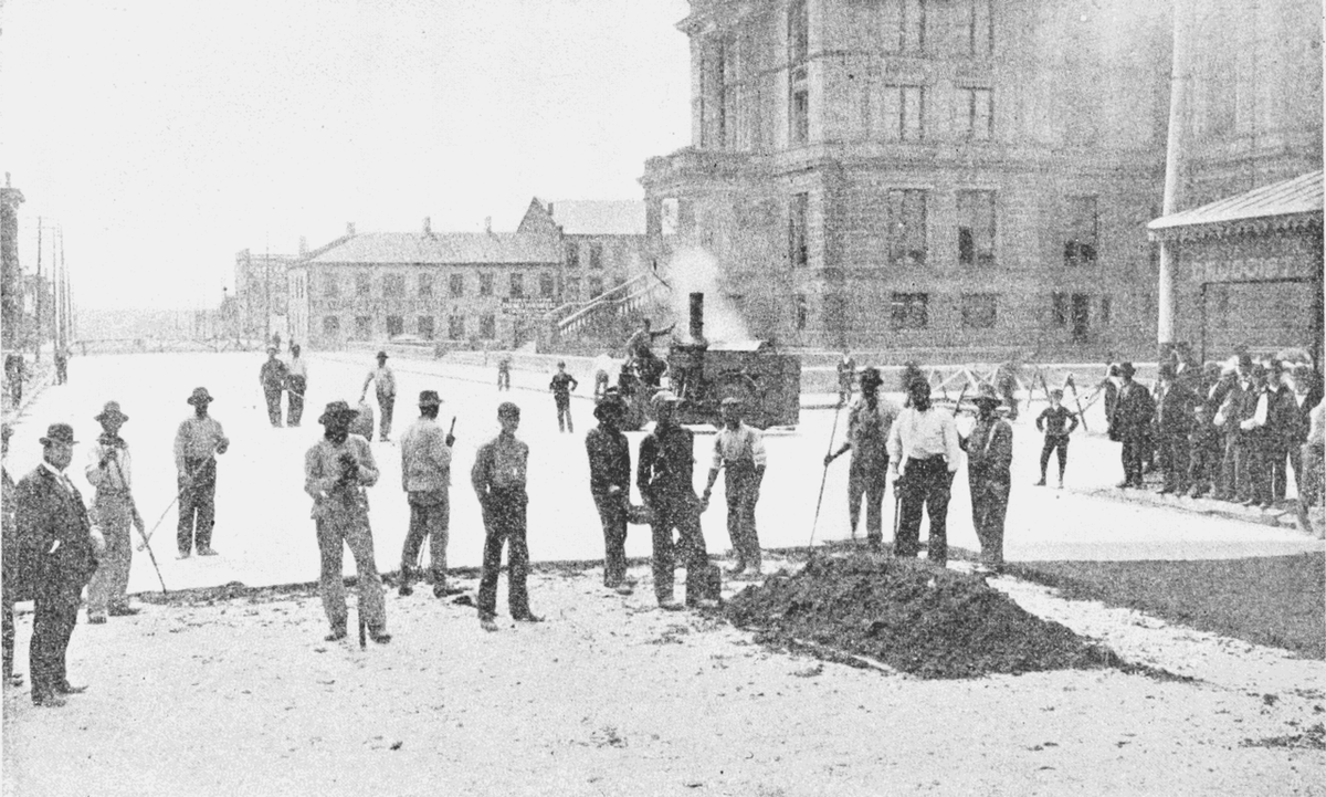 Arbeiter asphaltieren einen Platz. Hinten ist eine bereits asphaltierte, glatte Fläche. Im Vordergrund wird noch asphaltiert: Die meisten Arbeiter halten Geräte in der Hand. © Wikimedia