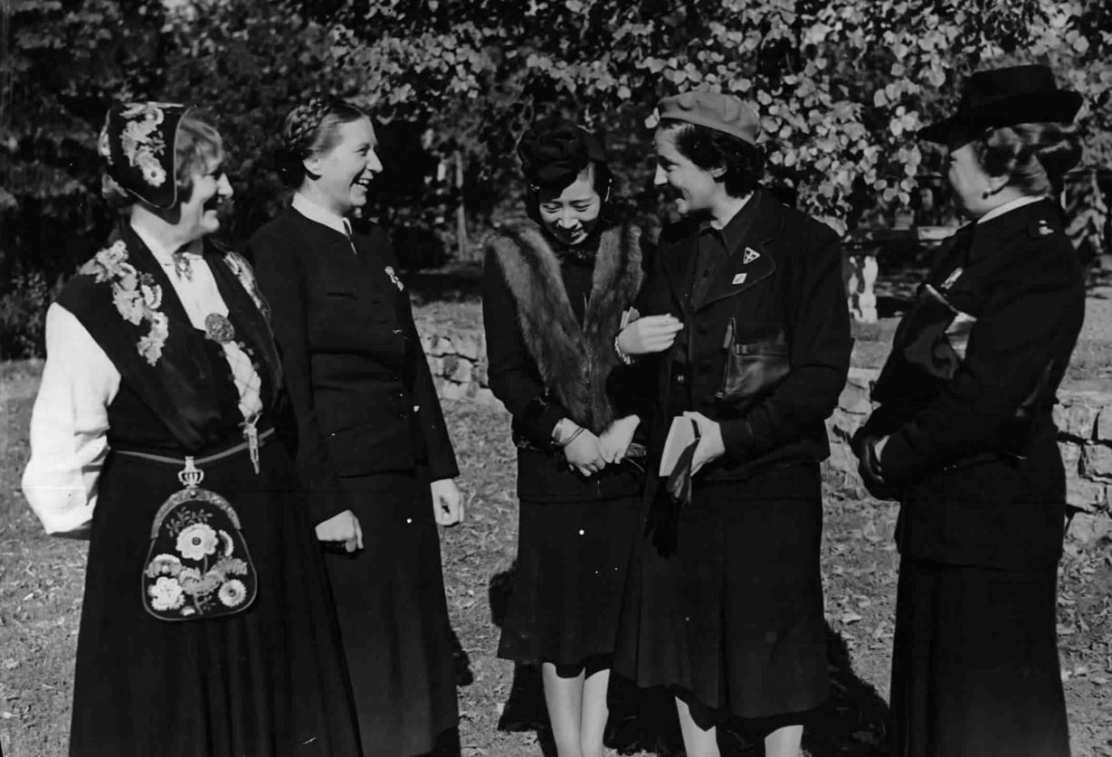 nf Frauen in dunklen Mänteln stehen in einem Park oder Garten beieinander, lachen und reden. Links steht die norwegische Frauenführerin Bjoner in Tracht mit Blumenmustern, rechts davon die Reichsfrauenführerin Scholtz-Klink mit einer Flechtfrisur, in der Mitte Frau Oshima, Gattin des japanischen Botschafters, mit Pelz und rechts die spanische Frauenführerin Primo de Rivera und die Italienerin Olga Medici, beide mit Hut.  © ÖNB Bildarchiv, Treffen faschistischer Frauen in Berlin, 1941. 