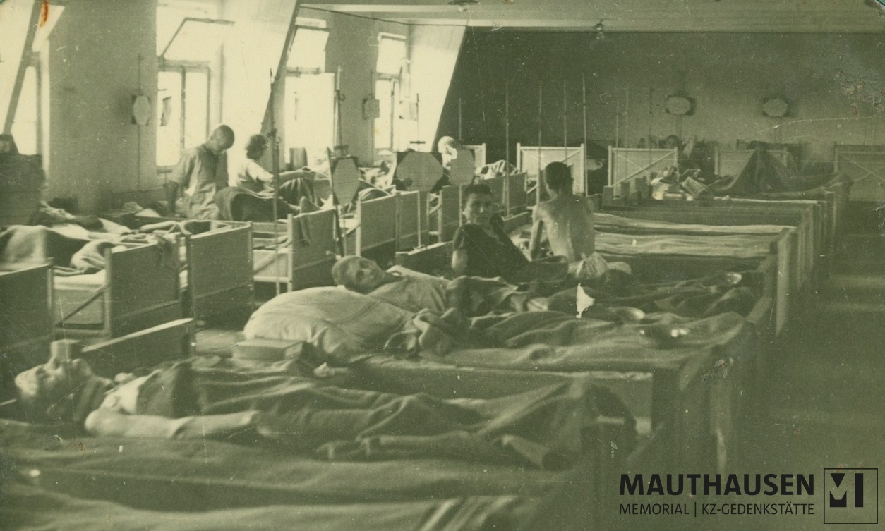 Das Schwarz-Wei&szlig;-Foto zeigt Bettenreihen in einem Innenraum. In den Betten liegen und sitzen geschw&auml;chte M&auml;nner. Zwei der ehemaligen H&auml;ftlinge schauen in die Kamera. &copy; Mauthausen Memorial, Fotoalbum Jerzy Ginter, Sanit&auml;tsbereich der US-Armee im befreiten KZ Gusen, Mai/Juni 1945, Signatur: 4.4.2.1.013.