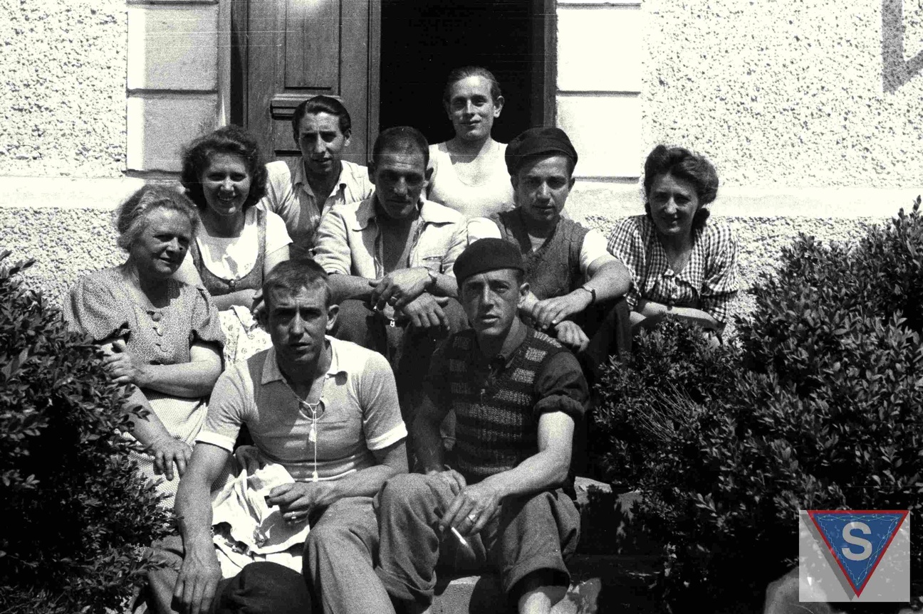 Das Schwarz-Wei&szlig;-Foto zeigt eine Gruppe von Menschen, die vor einem Haus auf der Treppe zur Eingangst&uuml;r sitzen. Ganz links sitzt Anna Pointner. Sie tr&auml;gt eine Bluse mit Kn&ouml;pfen und kurzen &Auml;rmeln. Ihre Haare sind hochgesteckt. In der Mitte der Gruppe sitzen sechs Spanier, die f&uuml;r die Kamera posieren. Einer davon l&auml;chelt, die anderen schauen ernst in die Kamera. Ein Mann ganz vorne h&auml;lt eine Zigarette in der Hand. Links und rechts der Spanier sitzt jeweils eine Tochter von Anna Pointner und l&auml;chelt in die Kamera. Im Vordergrund rahmen B&uuml;sche die Gruppe. &copy; Museu d'Hist&ograve;ria de Catalunya, Fons Amical de Mauthausen, Fotograf: Francisco Boix, Anna Pointner mit zwei T&ouml;chtern und sechs Spaniern vor ihrem Haus in Mauthausen, Mai 1945. 