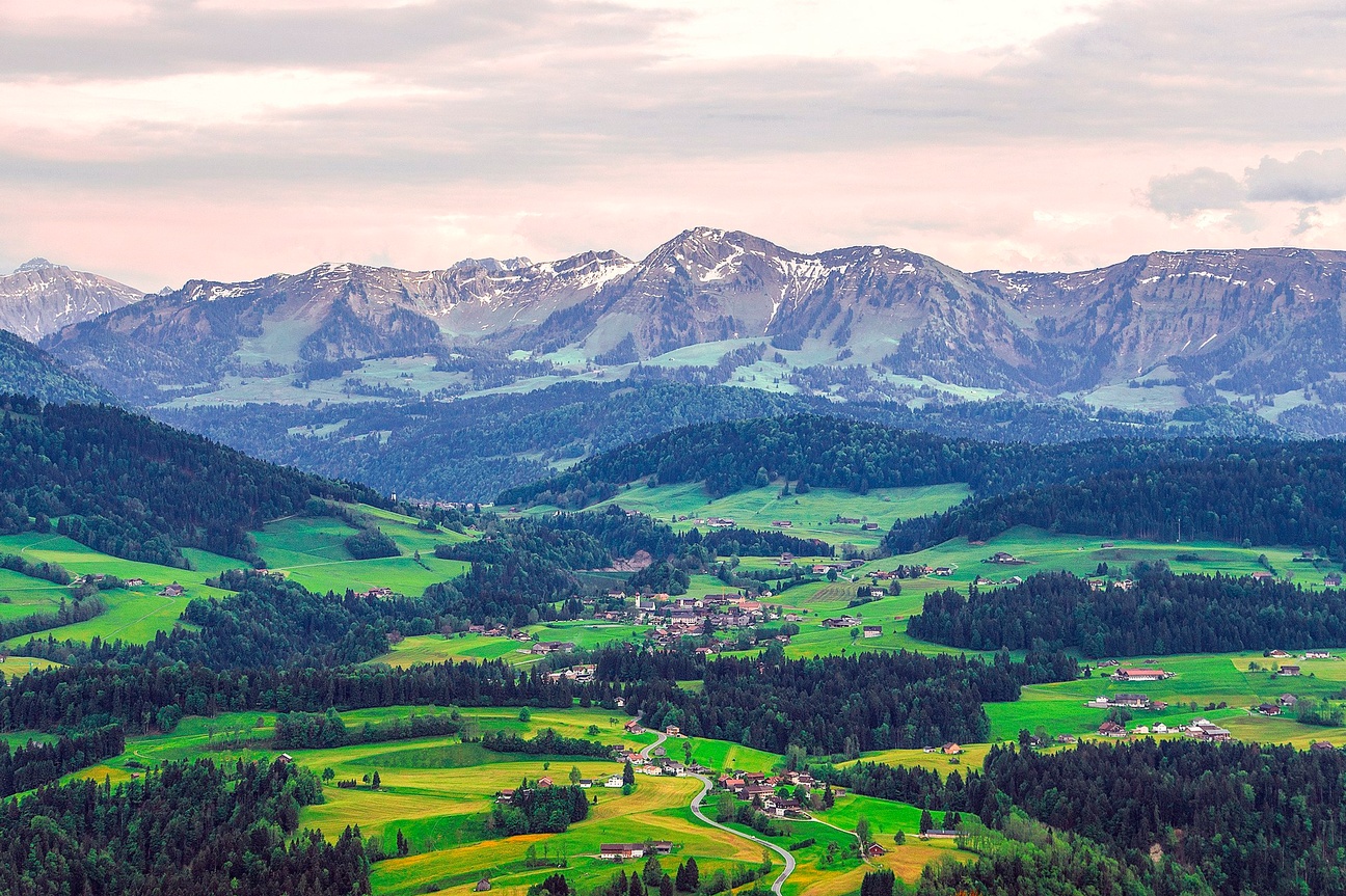 Blick auf das Dorf Krumbach: Eine Straße schlängelt sich durch die Landschaft. Zwischen gelben und hellgrünen Feldern und dunkelgrünen Waldstücken befinden sich Häusergruppen und Bauernhöfe. Im Hintergrund befindet sich eine Bergkette mit Schneeresten.  © Wikimedia, P&M Fotografie Stefan Emmer, Krumbach, 2018 (CC-BY-SA-4.0). 