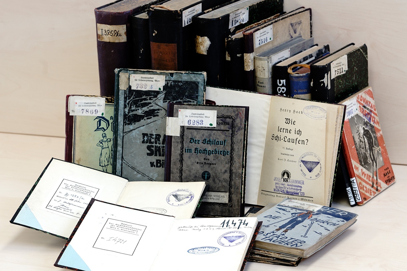 Alte Bücher mit zum Teil aufgeriebenen Kanten und Inventarnummern am Buchdeckel sind aufgereiht. Zwei Bücher im Vordergrund sind aufgeschlagen, Stempel sind sichtbar. Ein Titel lautet „Der Schilauf im Hochgebirge“, ein anderer „Wie lerne ich Schi-Laufen?“ © Marc Drews. 