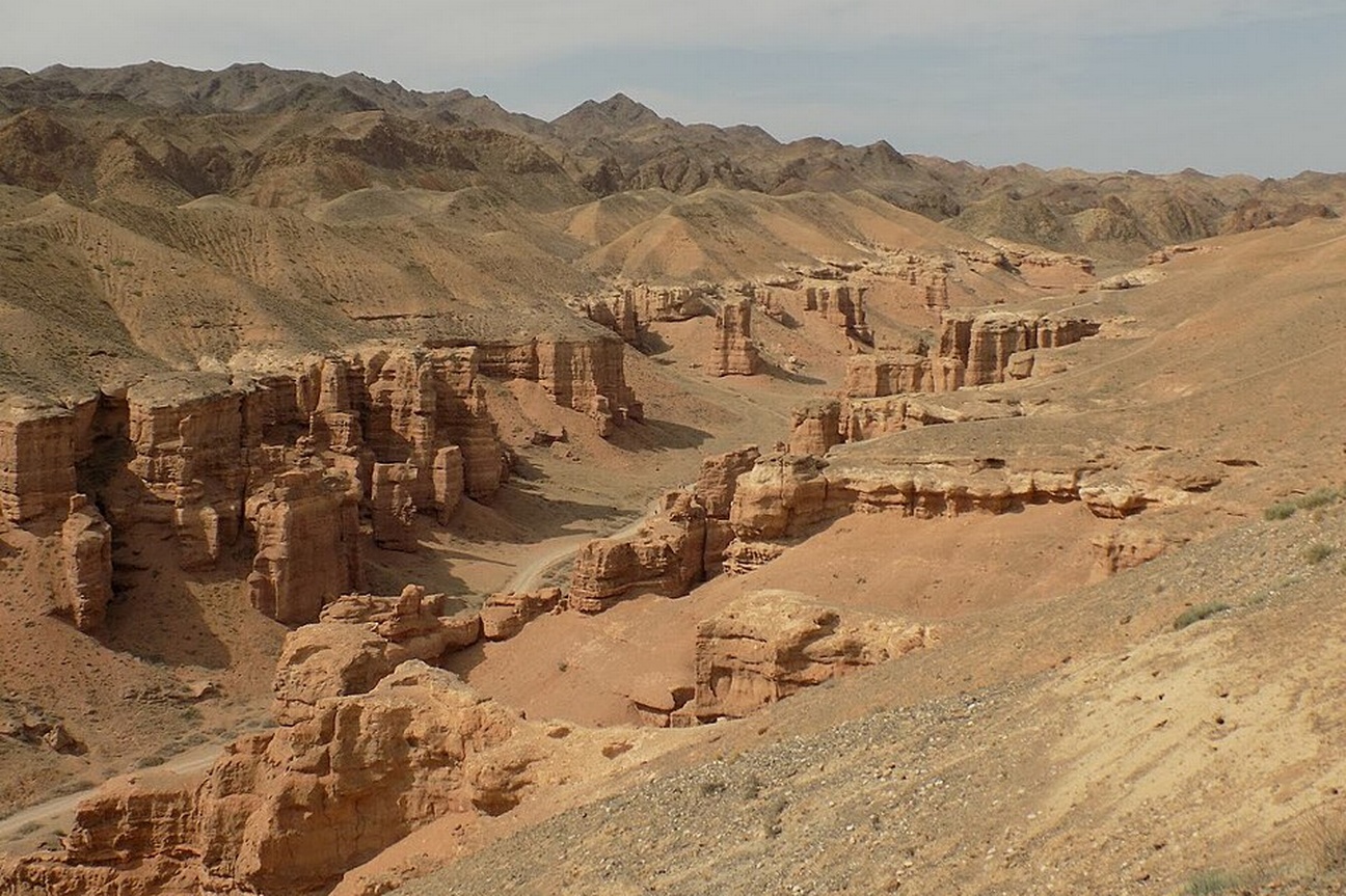 Das Foto zeigt einen Canyon mit Bergen im Hintergrund vor einem graublauen Himmel. Die Erde und das Gestein des Canyons sind orange.  © Wikimedia, Anna Dobrynina, Canyon in Kazakhstan, 2017 (CC-BY-3.0).  