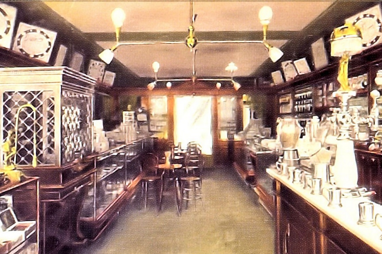 Das Bild zeigt das Innere einer Apotheke. In der Mitte befindet sich ein Gang mit Tischen und Stühlen. An den Wänden rechts und links stehen deckenhohe Schränke, in denen sich Gefäße befinden. Davor stehen lange Theken.   © Wikimedia, Apotheke in den USA, 1920. 