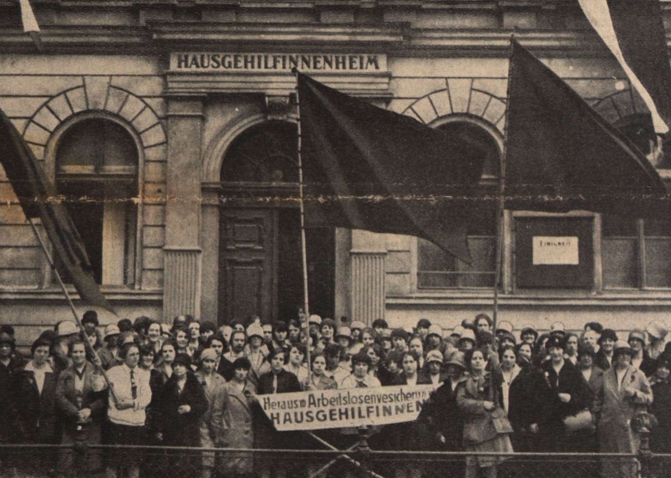 Eine große Gruppe Frauen in Festkleidung mit drei großen Fahnen und einem Banner steht vor dem Eingang zu einem Haus. Auf dem Banner ist zu lesen: Heraus mit der Arbeitslosenversicherung für Hausgehilfinnen.  © ANNO ÖNB