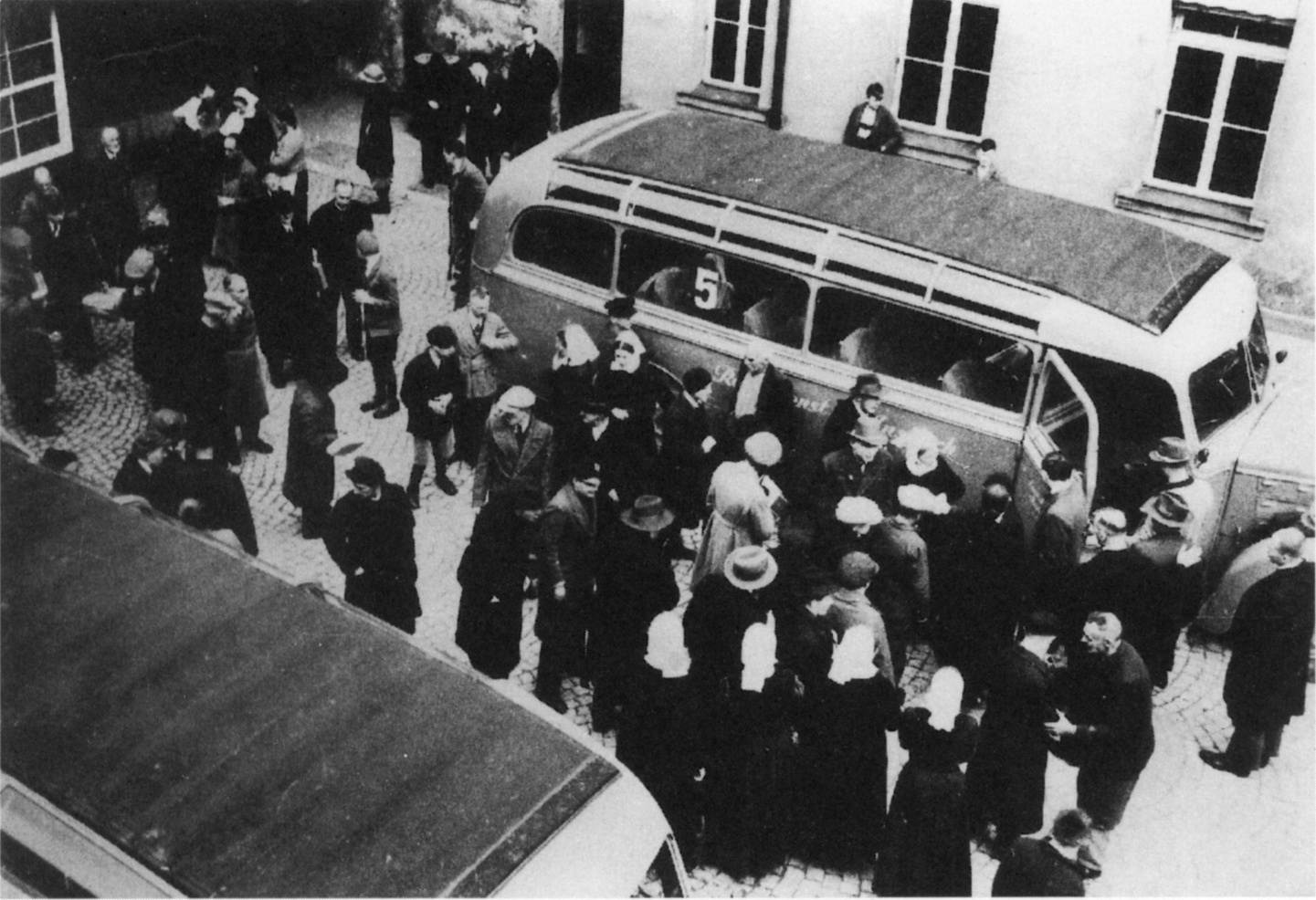 Auf einem gepflasterten Platz umgeben von Gebäuden stehen zwei Busse. Zwischen den leeren Bussen stehen mehrere Dutzend Menschen. © Wikimedia, Pressestelle der Diakonie Neuendettelsau, Aktion T4 (Diakonie Neuendettelsau), 1941