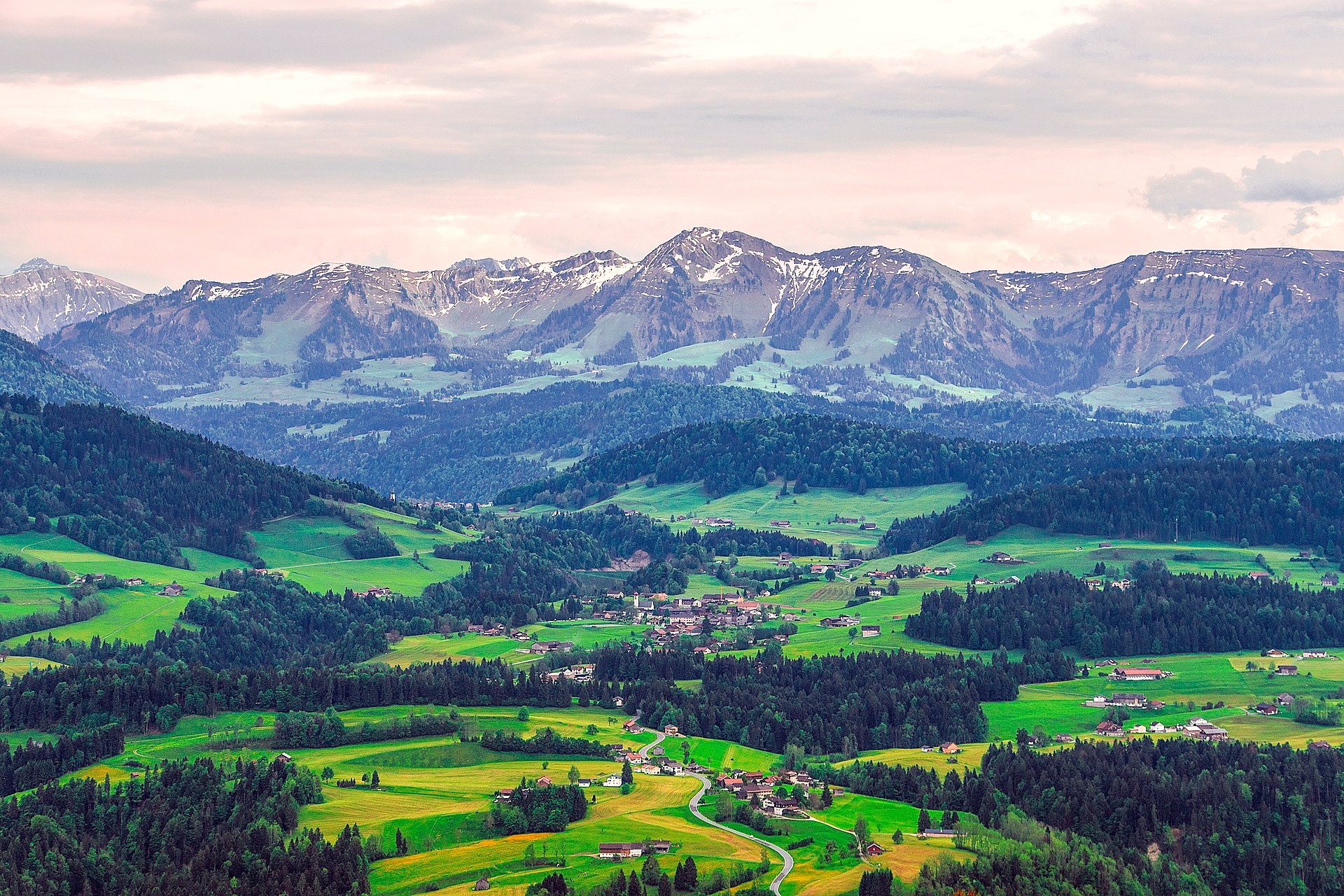 Blick auf das Dorf Krumbach: Eine Straße schlängelt sich durch die Landschaft. Zwischen gelben und hellgrünen Feldern und dunkelgrünen Waldstücken befinden sich Häusergruppen und Bauernhöfe. Im Hintergrund befindet sich eine Bergkette mit Schneeresten.  © Wikimedia, P&M Fotografie Stefan Emmer, Krumbach, 2018 (CC-BY-SA-4.0). 