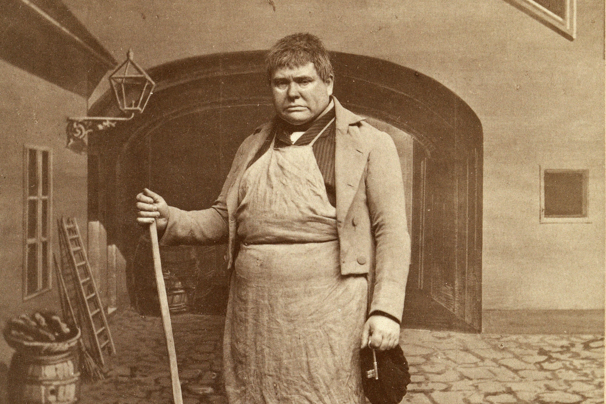 Ein Mann steht mit Besen und Schlüssel in der Hand auf einem gepflasterten Platz. Hinter ihm ist ein Tor zu sehen.  © Wien Museum